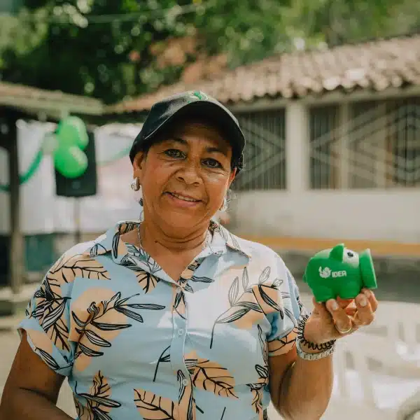 Un plano medio horizontal en exteriores con buena luz solar muestra a una mujer de tez morena y mediana edad, de pie, mirando y sonriendo a la cámara  Lleva una gorra de béisbol negra con un pequeño logo verde en el centro, pendientes colgantes plateados, un collar de cadena plateada fina y una camisa de botones de manga corta azul claro con un estampado de hojas beige y marrón claro  Sostiene una pequeña alcancía de plástico verde con el logo blanco de IDEA hacia arriba con la mano izquierda a la altura del hombro, que se curva hacia arriba en la parte inferior  La alcancía parece una mezcla de una alcancía tradicional y una bombilla de silicona, con una abertura redonda y ancha en el extremo donde estarían el morro y la boca  El fondo está ligeramente desenfocado, con la luz del sol asomando por detrás de un árbol verde frondoso en la parte superior izquierda, una pancarta con globos verdes en la parte superior izquierda, una casa de color blanco cremoso con tejas de arcilla marrón en el centro del fondo, y un grupo de sillas de plástico blanco apiladas en la parte inferior derecha  La luz es brillante y clara, pero no dura 