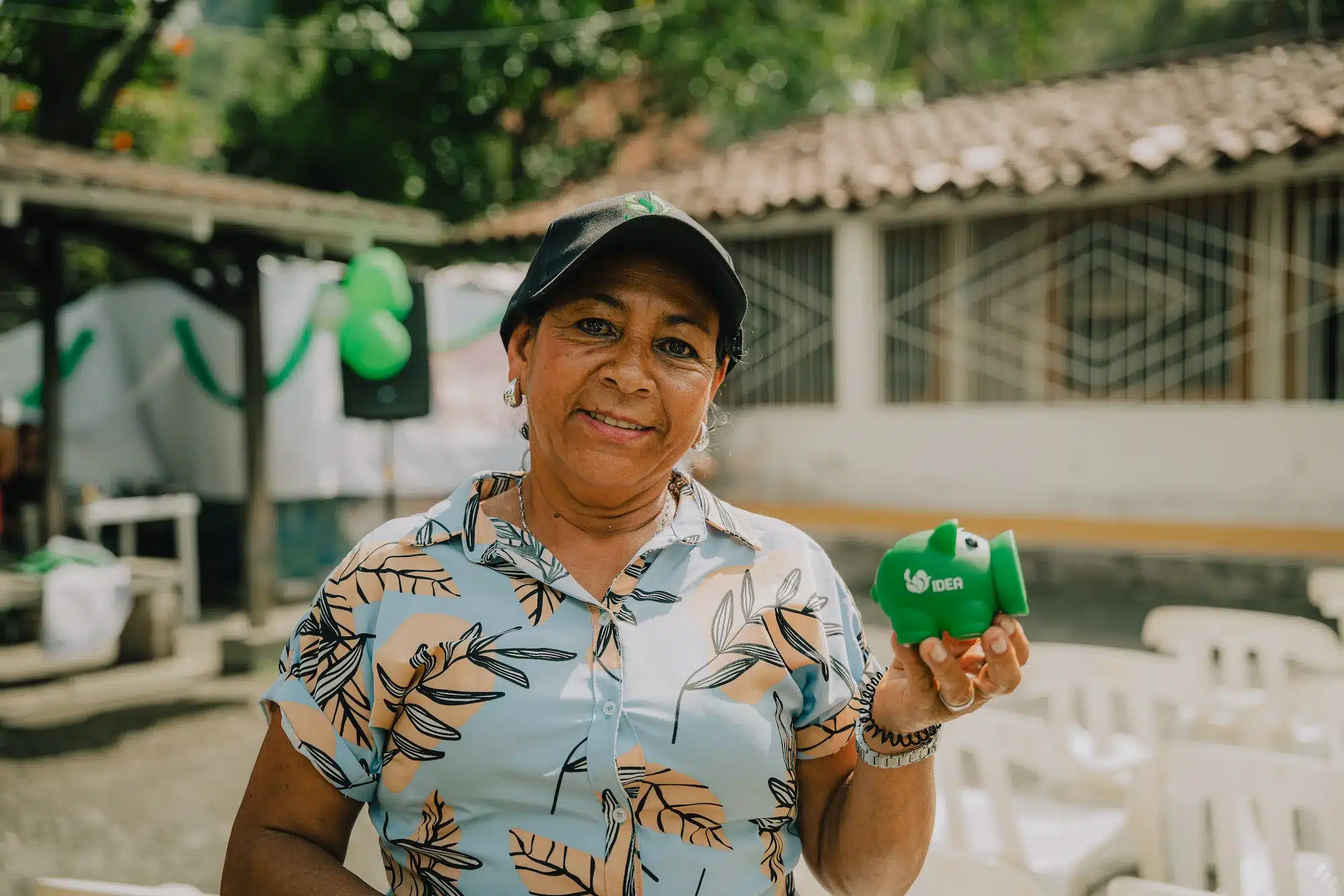 Un plano medio horizontal en exteriores con buena luz solar muestra a una mujer de tez morena y mediana edad, de pie, mirando y sonriendo a la cámara. Lleva una gorra de béisbol negra con un pequeño logo verde en el centro, pendientes colgantes plateados, un collar de cadena plateada fina y una camisa de botones de manga corta azul claro con un estampado de hojas beige y marrón claro. Sostiene una pequeña alcancía de plástico verde con el logo blanco de IDEA hacia arriba con la mano izquierda a la altura del hombro, que se curva hacia arriba en la parte inferior. La alcancía parece una mezcla de una alcancía tradicional y una bombilla de silicona, con una abertura redonda y ancha en el extremo donde estarían el morro y la boca. El fondo está ligeramente desenfocado, con la luz del sol asomando por detrás de un árbol verde frondoso en la parte superior izquierda, una pancarta con globos verdes en la parte superior izquierda, una casa de color blanco cremoso con tejas de arcilla marrón en el centro del fondo, y un grupo de sillas de plástico blanco apiladas en la parte inferior derecha. La luz es brillante y clara, pero no dura.